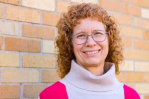 headshot of Cheri with red hair, glasses, and a turtle neck sweater that is cream with a red block on shoulder going all the way down her side. She is standing near a brick wall on a sunny day.